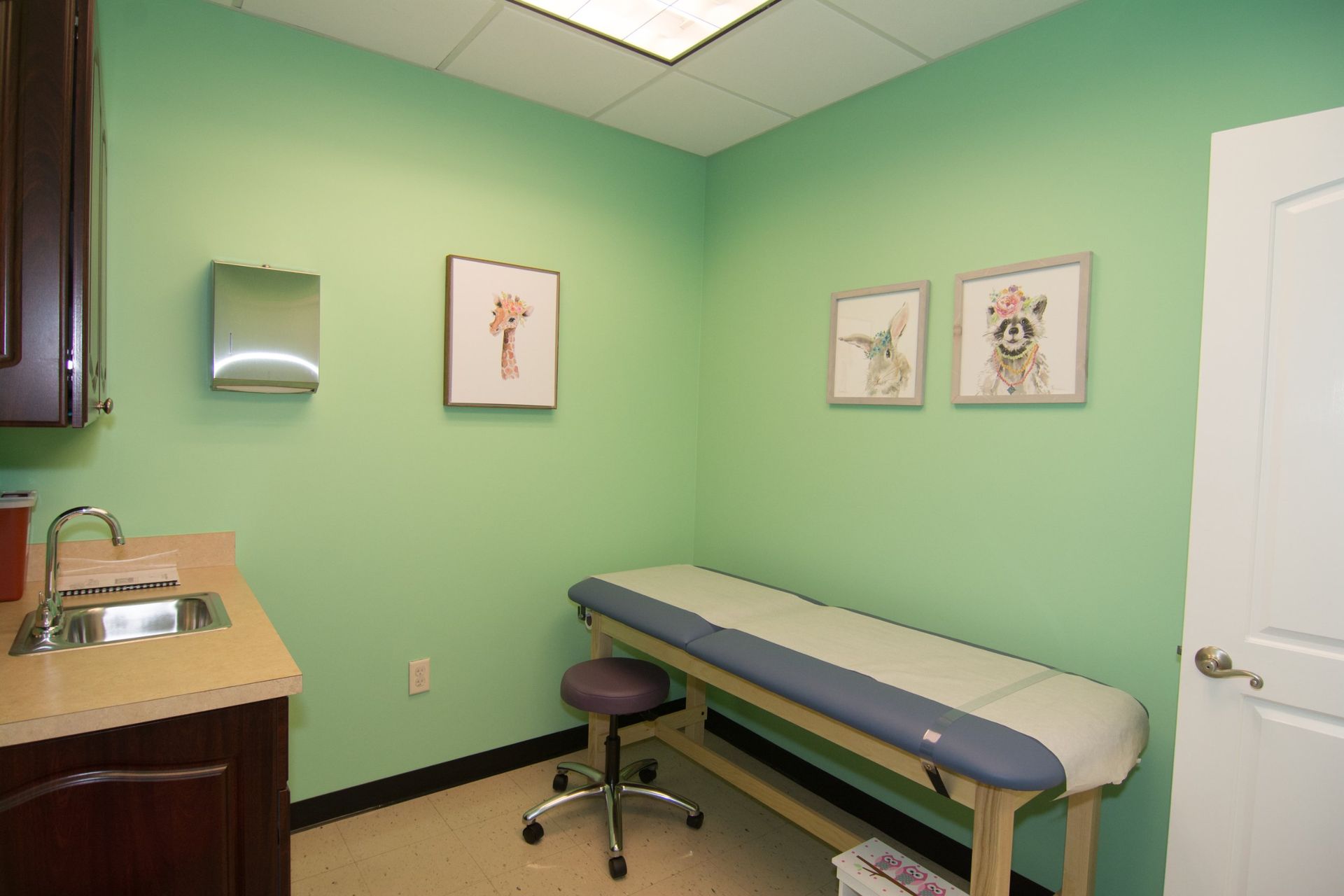 photo of the consultation room with light green walls, one bed against the wall beside the white door on the right, a sink on the other side and three framed drawings hanging on the wall.