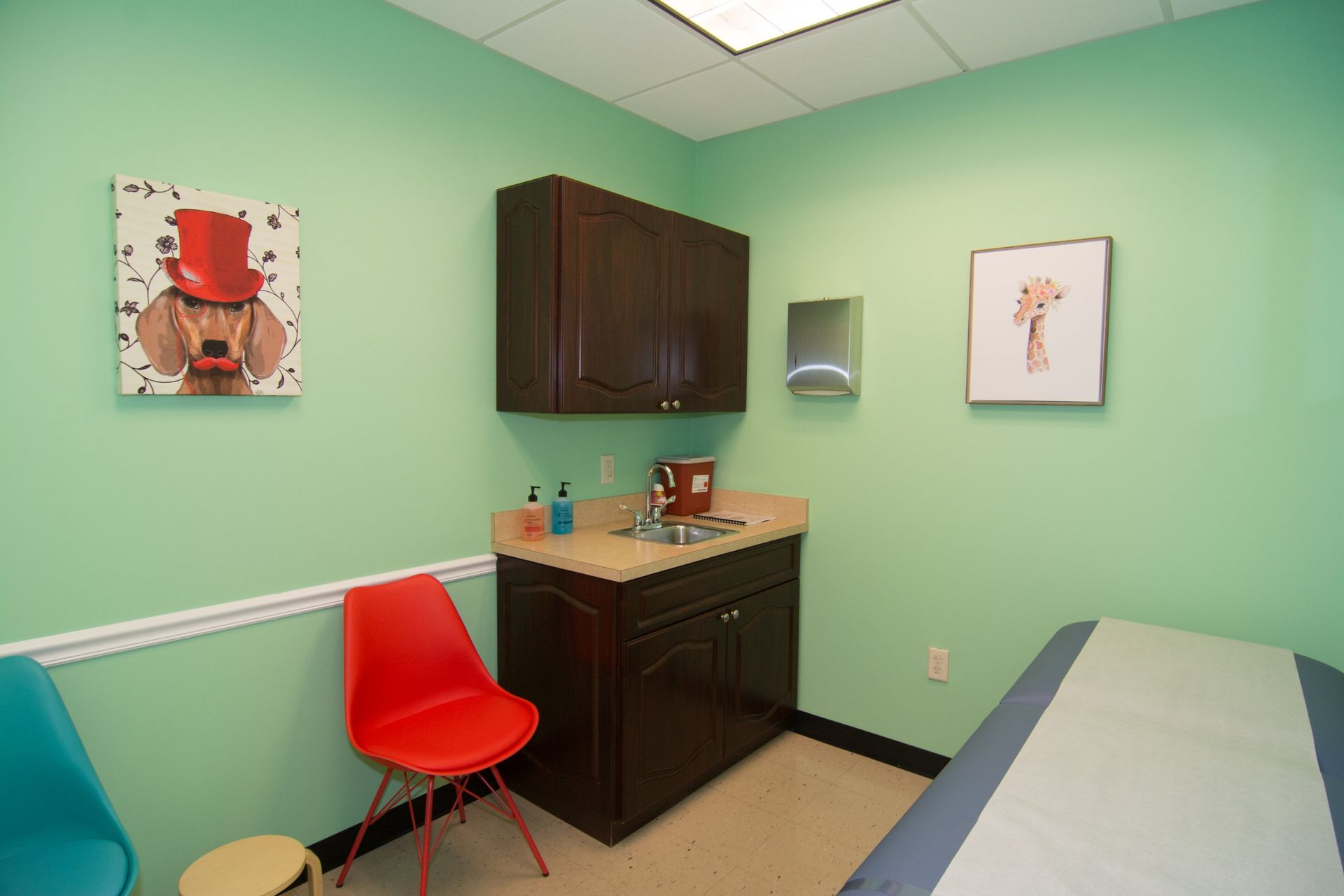 Photo of the consultation room with a bed, red and blue chair beside the sink and closet, green walls with one square painting hanging on each side.