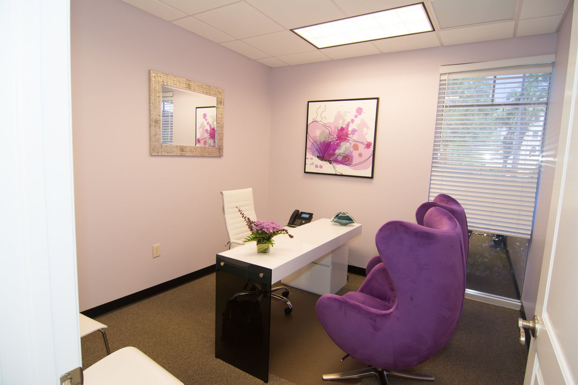 Photo inside the consultation office. White table, white swivel chair for the doctor, two purple for the patients.