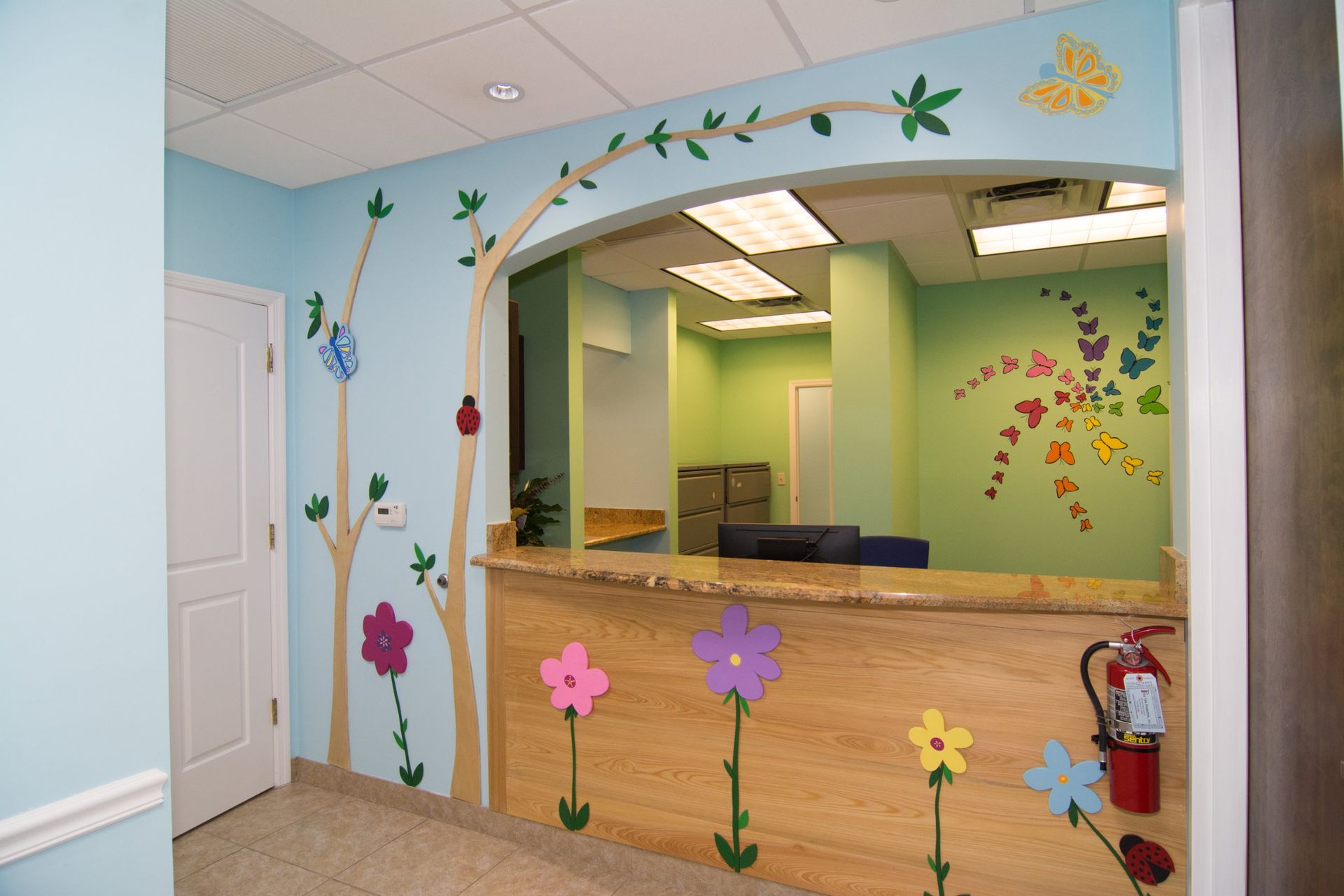 Photo of the front desk. Light Blue wall with flower decals and butterflies, wooden counter with nobody behind it, white door on the right.