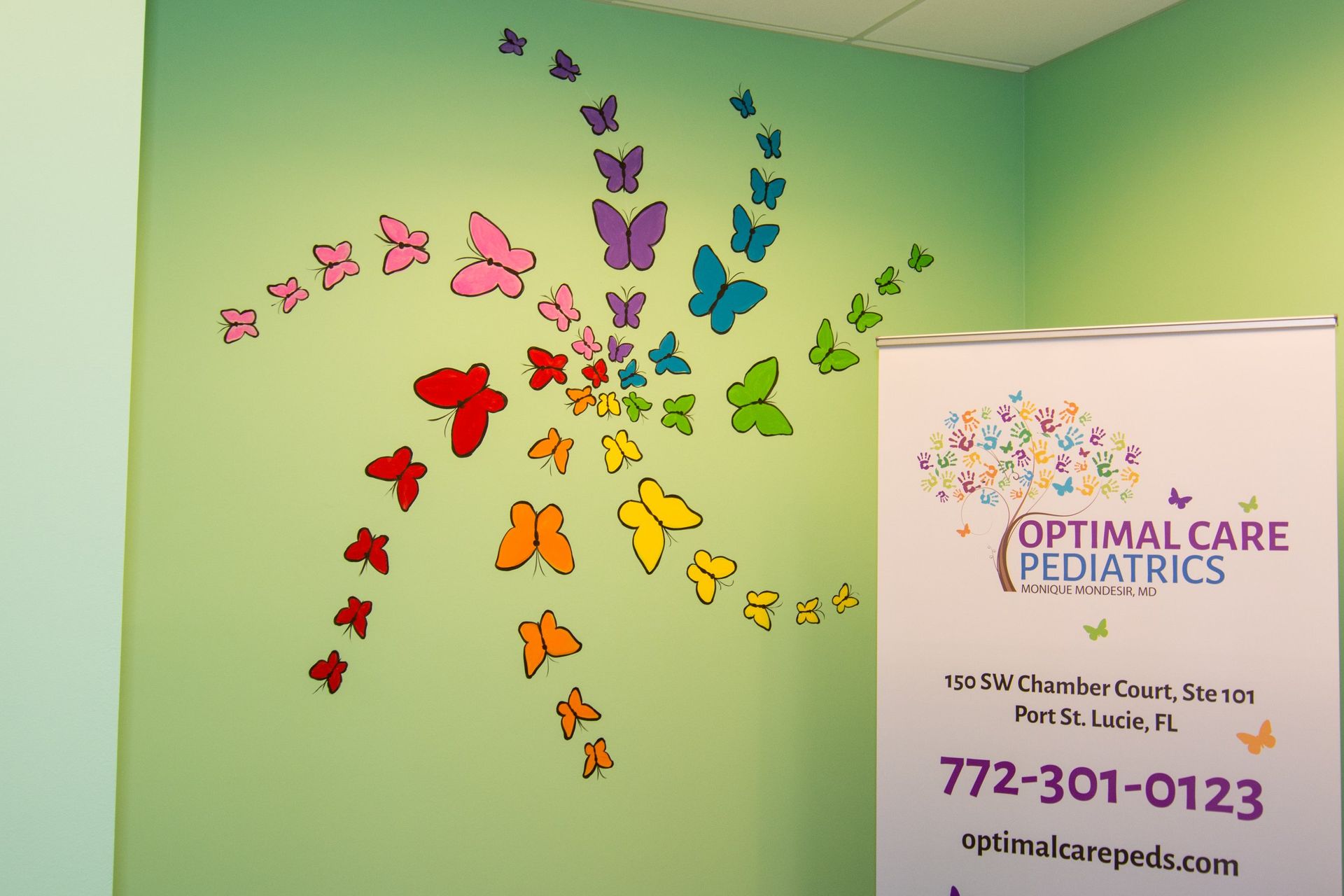 Spiralling butterflies stickers on a green wall, beside a tarp with Optimal Care Pediatrics logo, address, phone number and email 