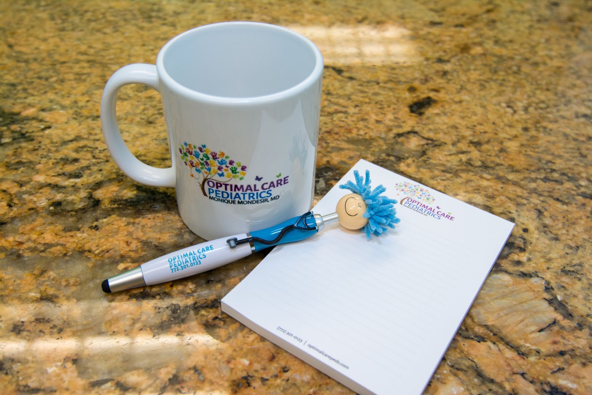 a blue and white pen with in between a mug and a pad with Optimal Care Pediatrics Logo