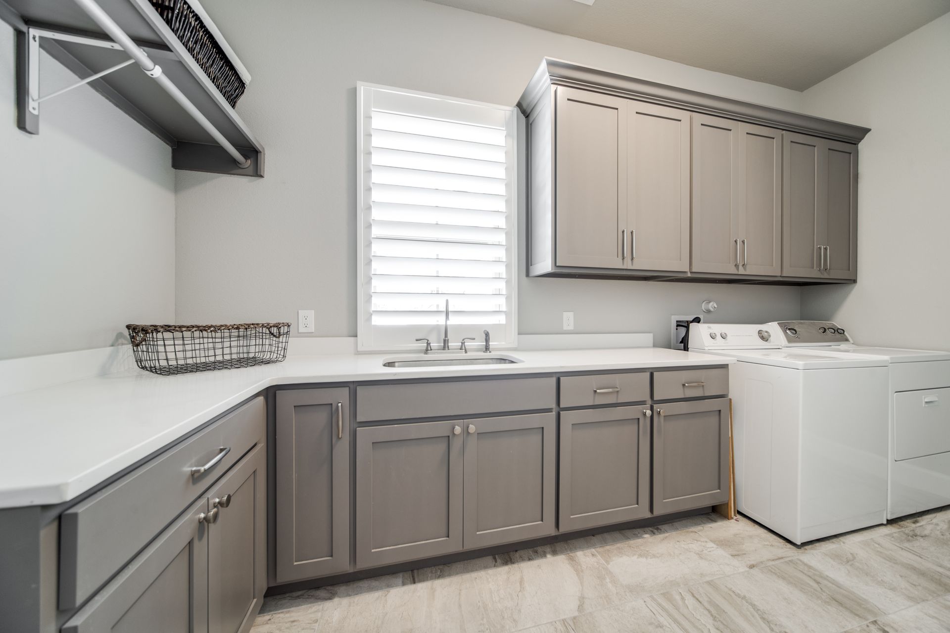 A laundry room with gray cabinets , white appliances , a sink and a window.