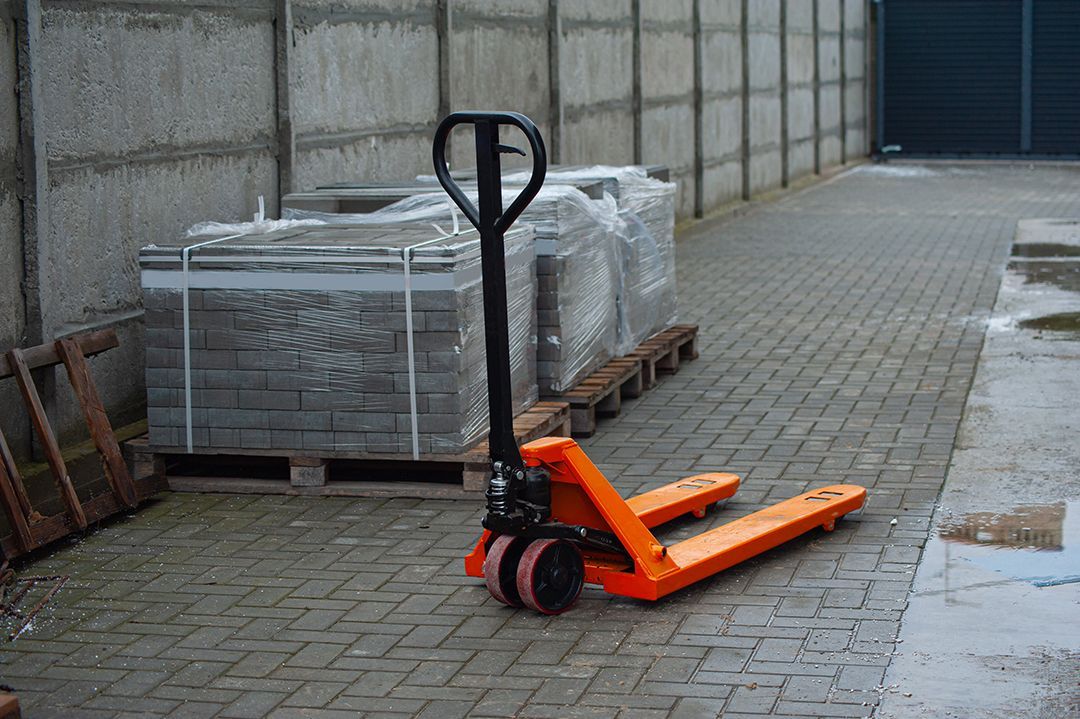 Una transpaleta naranja se encuentra sobre una superficie pavimentada junto a una pila de materiales de construcción paletizados.