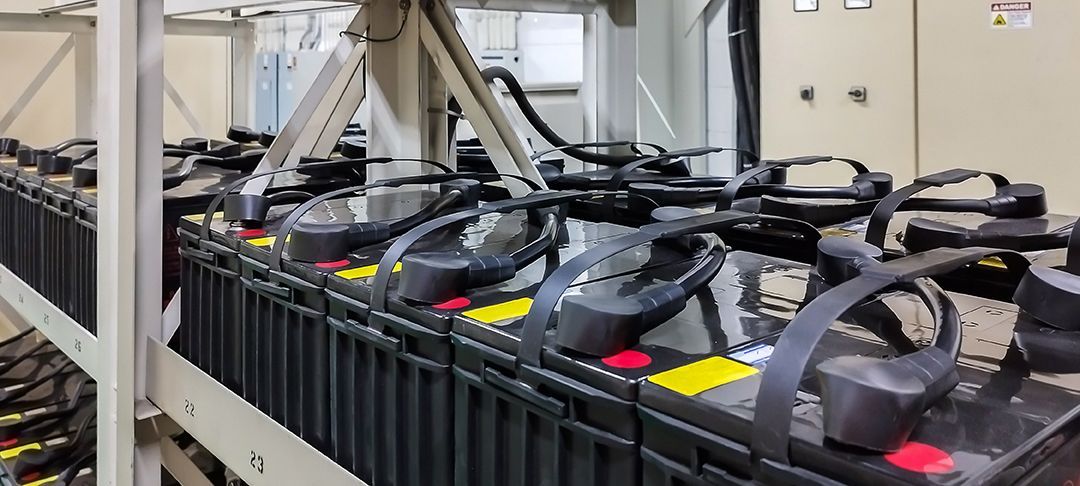 Filas de grandes baterías industriales negras almacenadas en estanterías metálicas, conectadas por cables eléctricos negros.