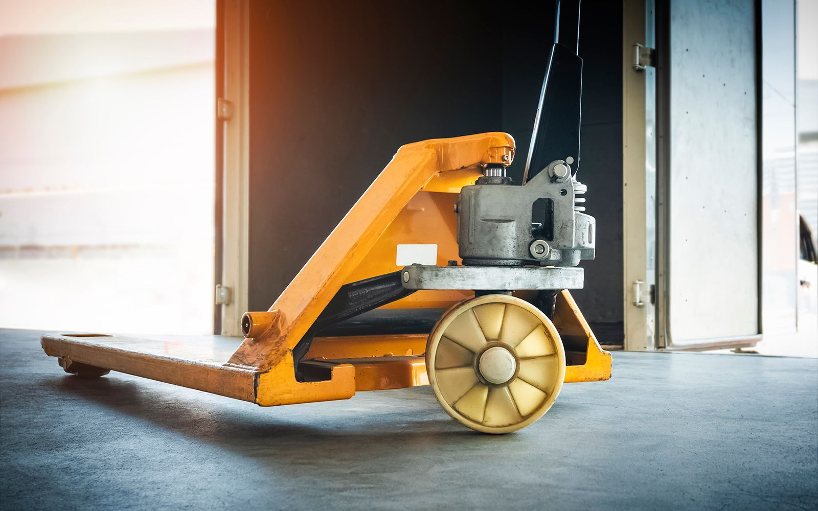Una transpaleta manual naranja está sobre un suelo de hormigón frente a una puerta abierta.