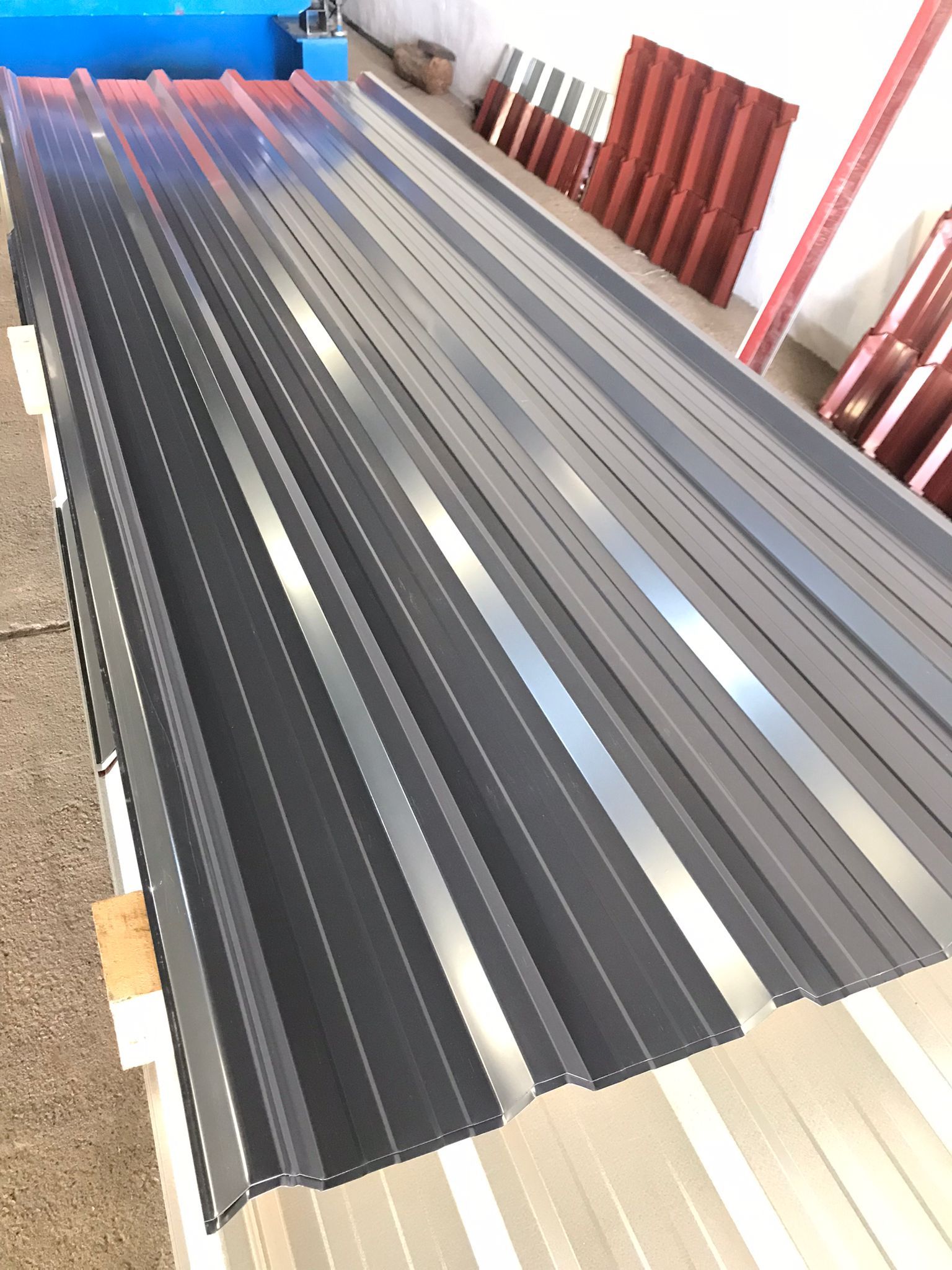 Láminas de techo de metal corrugado de color gris, apiladas con material de techo rojo en el fondo.