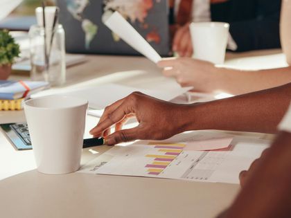 Mãos trabalhando em meio a papéis, gráficos e xícaras de café em uma mesa de reuniões.