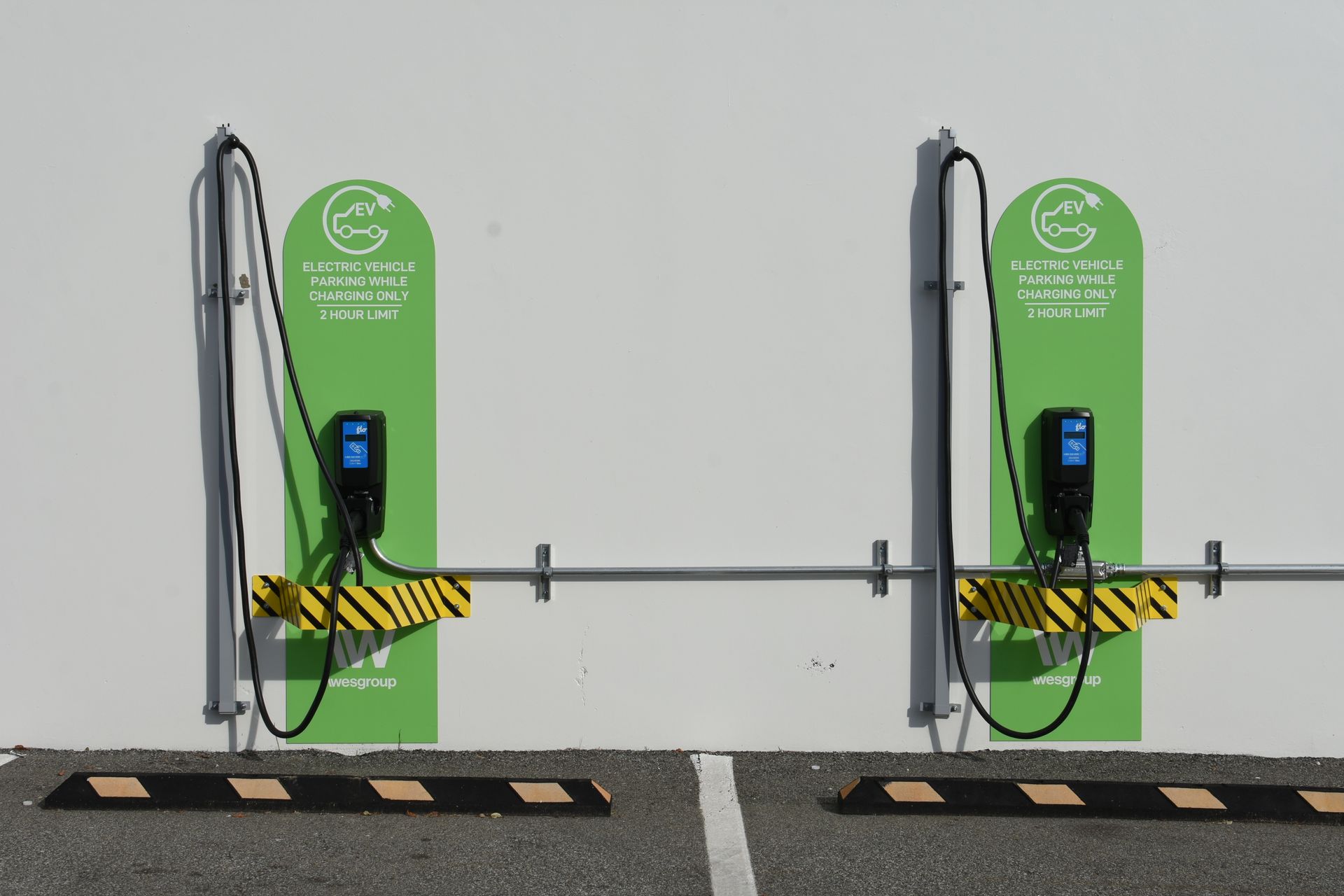 Two Wattz Up wall guards striped yellow and black are installed on the wall in front of two wall-mounted EV chargers.