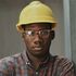 A man wearing a hard hat and glasses is leaning against a wall.