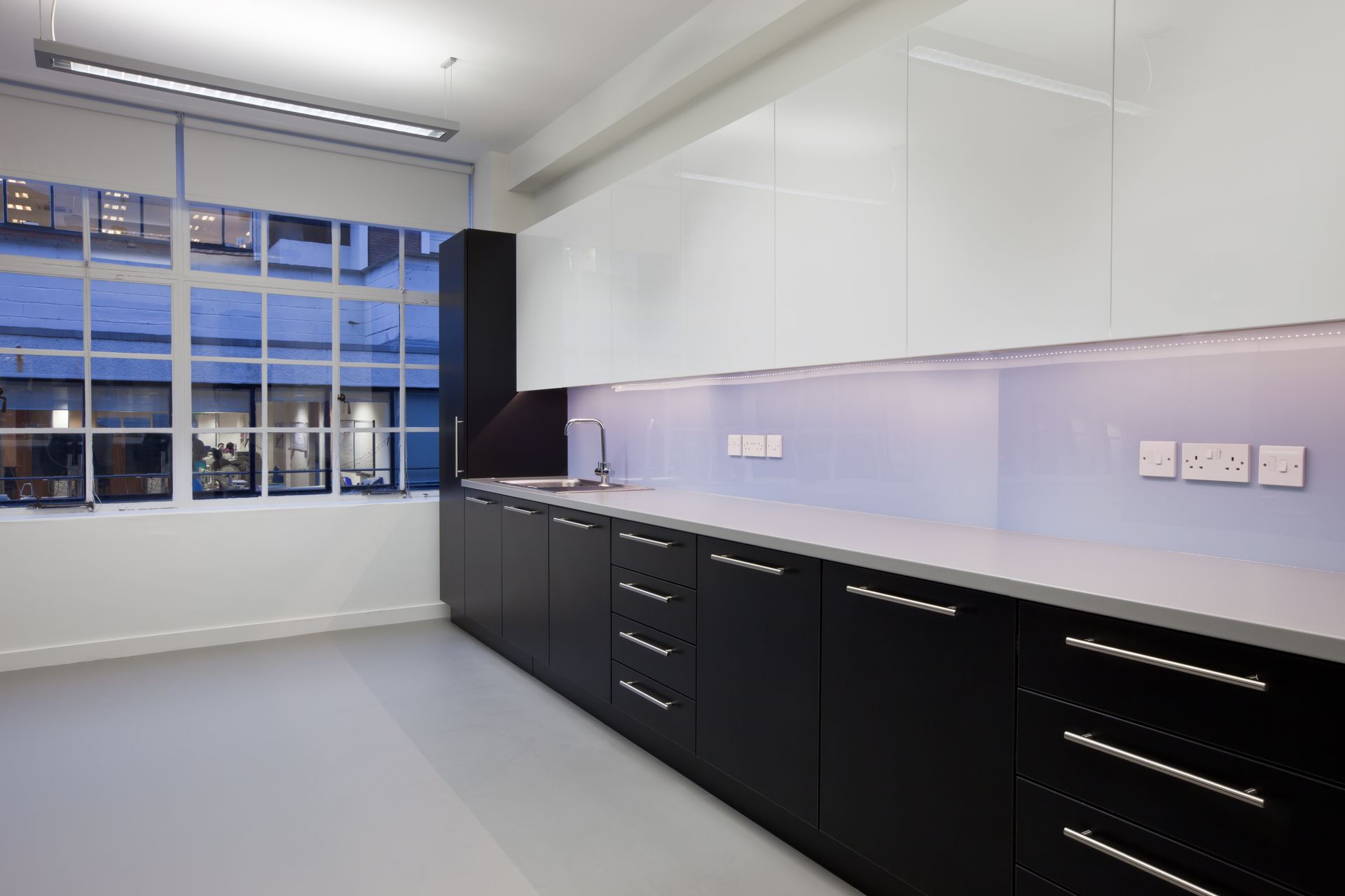 Modern office kitchen with black and white cabinets, window, sink, and fluorescent lighting.