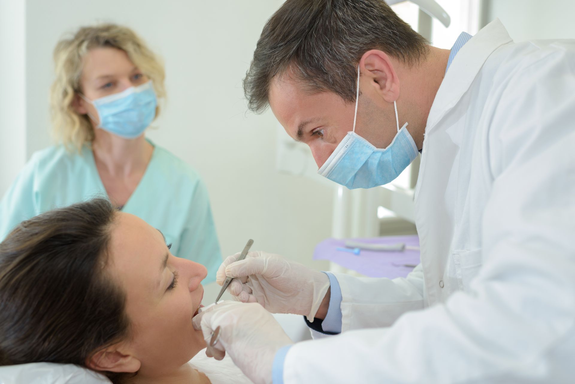 A dentist is inspecting the teeth of a patient. A dental assistant is in the background.
