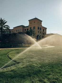 A sprinkler is spraying water on a lush green field in front of a large building.