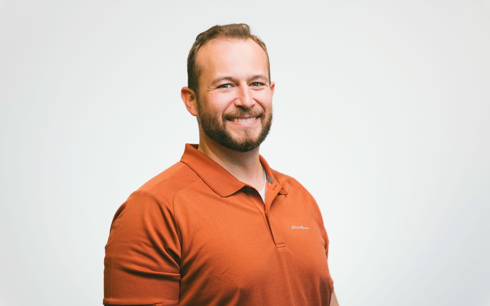 A man with a beard is wearing an orange polo shirt and smiling.