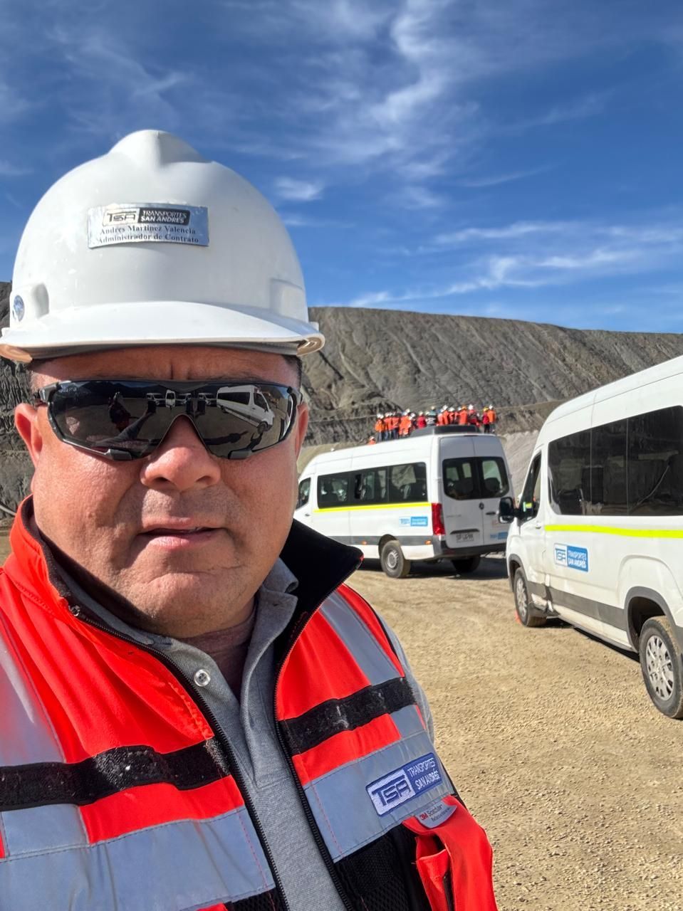 Hombre con equipo de seguridad, gafas de sol y casco, con furgonetas blancas en una cantera bajo un cielo azul.