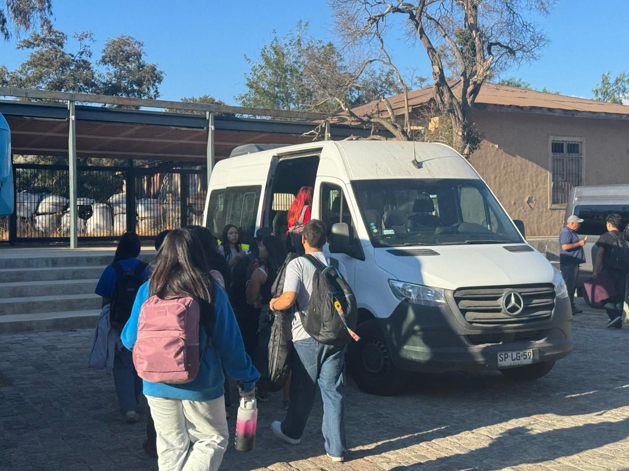 Estudiantes subiendo a una camioneta blanca frente a un edificio; algunos llevan mochilas. Día soleado.