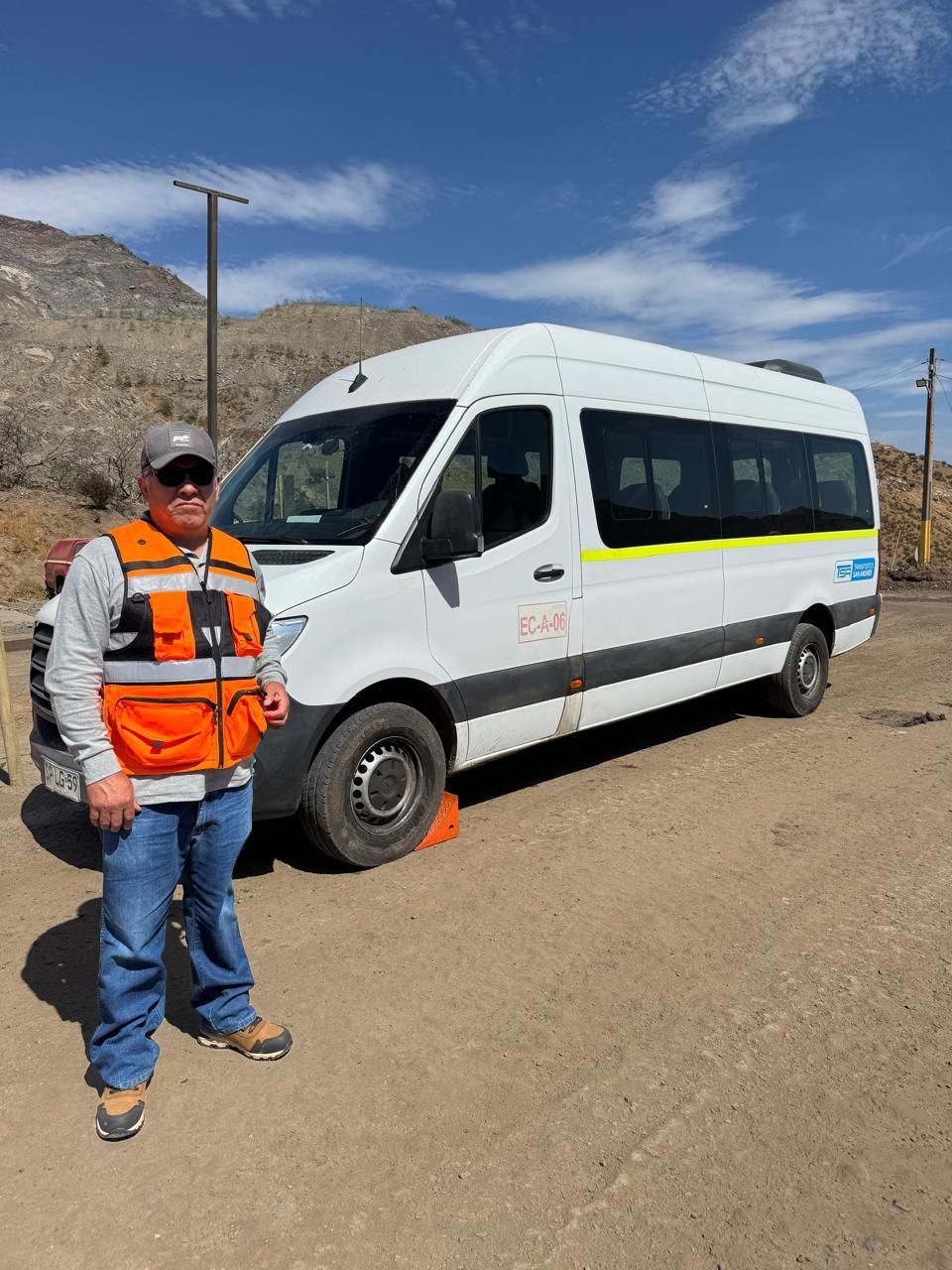 Un hombre con chaleco naranja se encuentra junto a una camioneta blanca en un camino de tierra bajo un cielo azul.