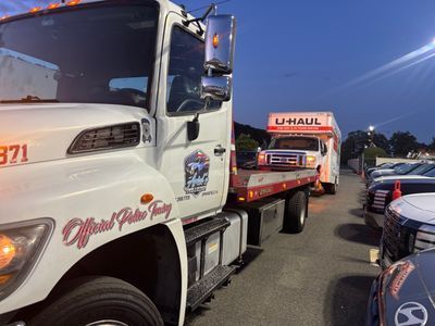Tow truck towing a U-Haul truck on a car lot; red, white, and blue colors; evening setting.