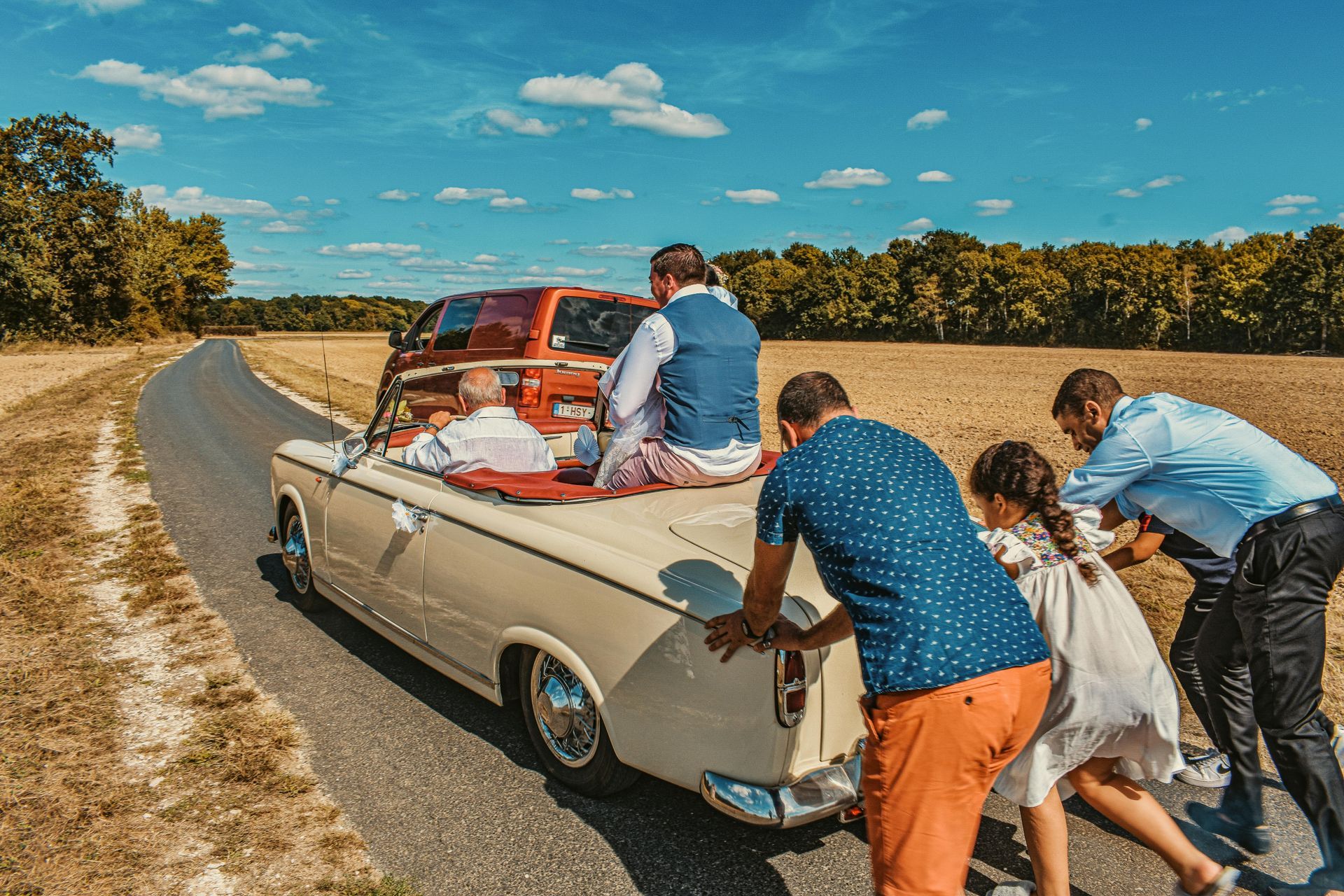 People pushing a vintage convertible on a rural road under a blue sky; someone sits in the car.