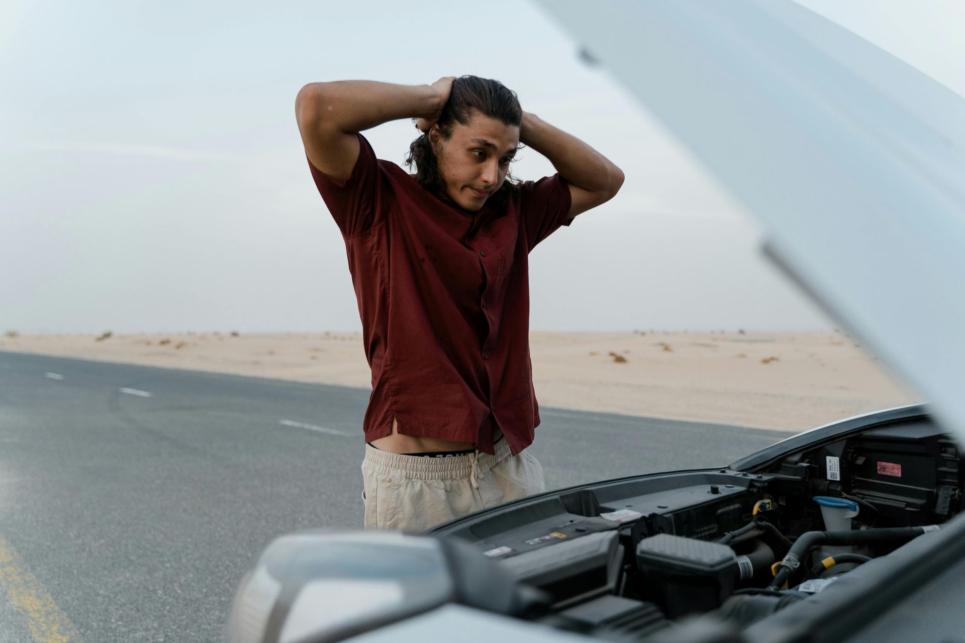 Man with hands on head, looking at open car hood on a desert road, appears stressed.