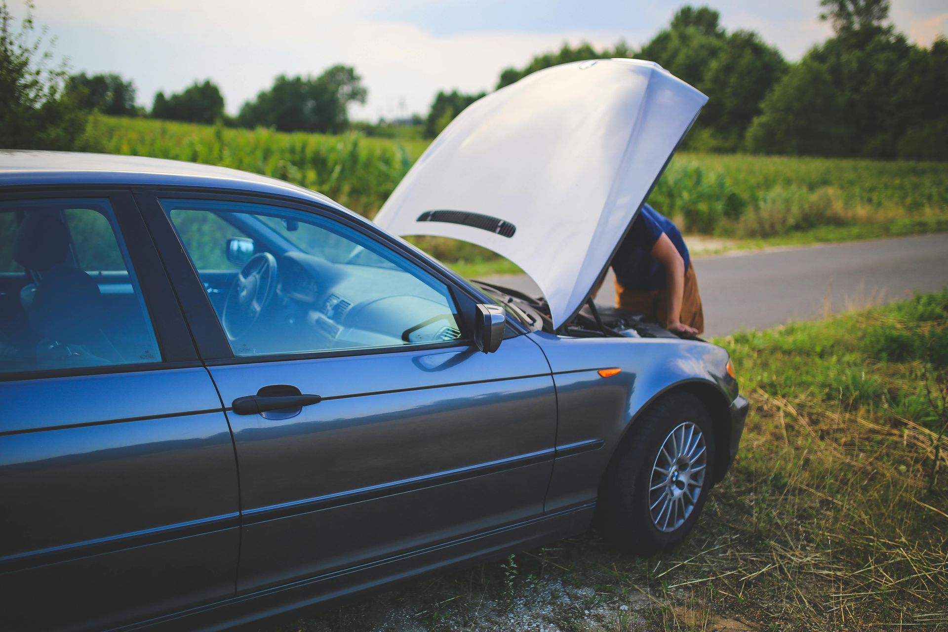 Person looking under the hood of a blue car on the side of a rural road.