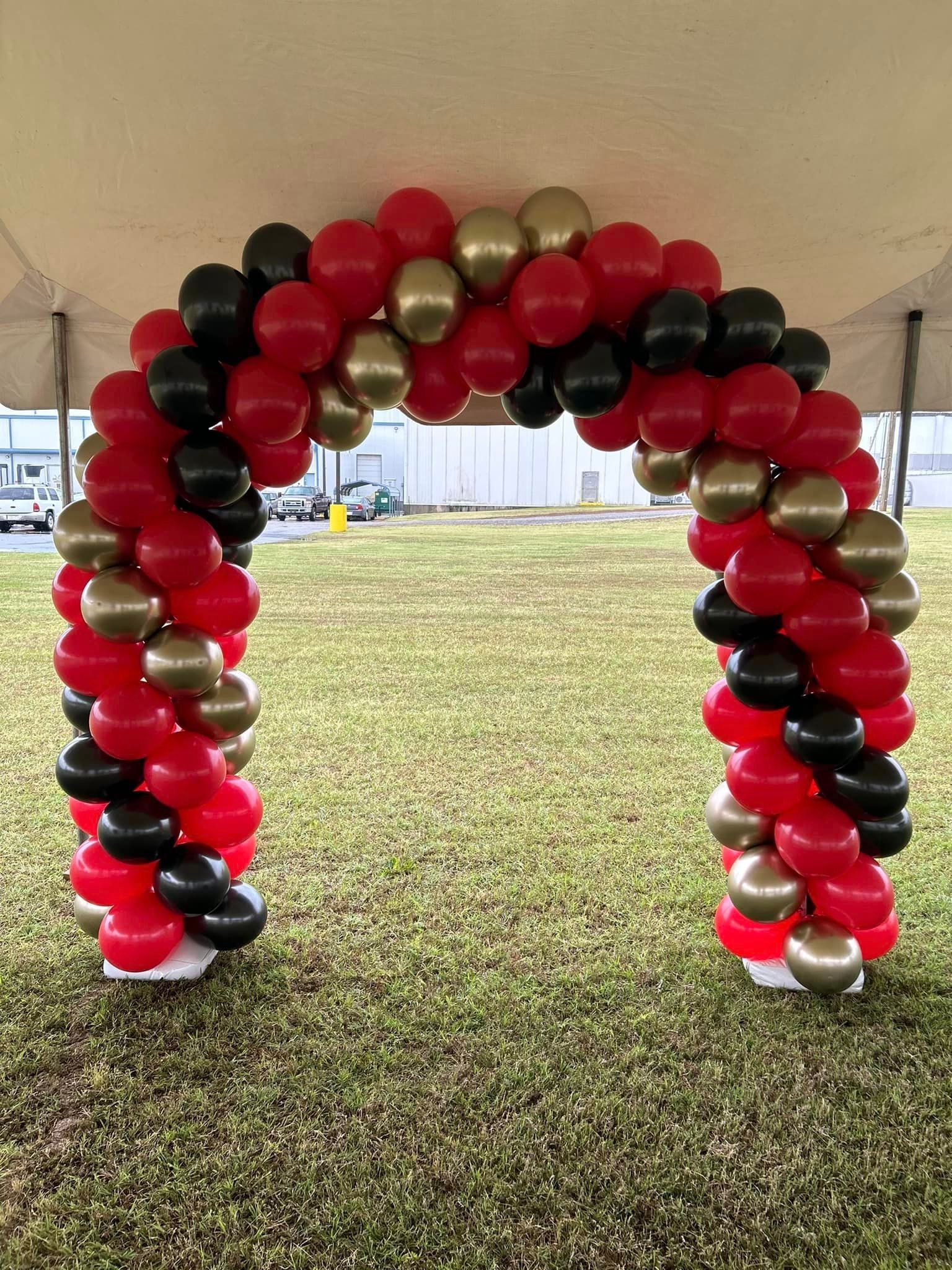 A red , black and gold balloon arch is sitting in the grass under a tent.