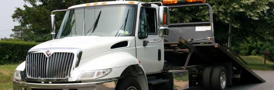 White tow truck with black bed parked on the road, with trees in the background.