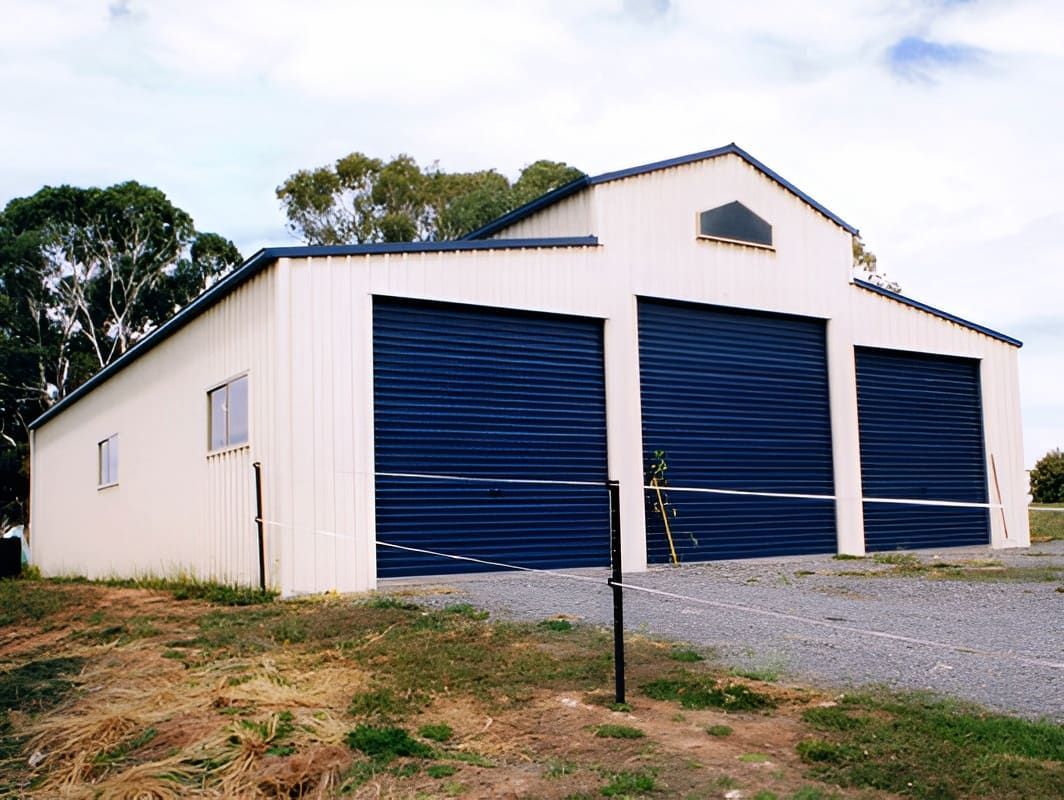 White Metal Building With Three Blue Garage Doors on a Gravel Driveway — Northern Rivers Sheds in South Lismore, NSW