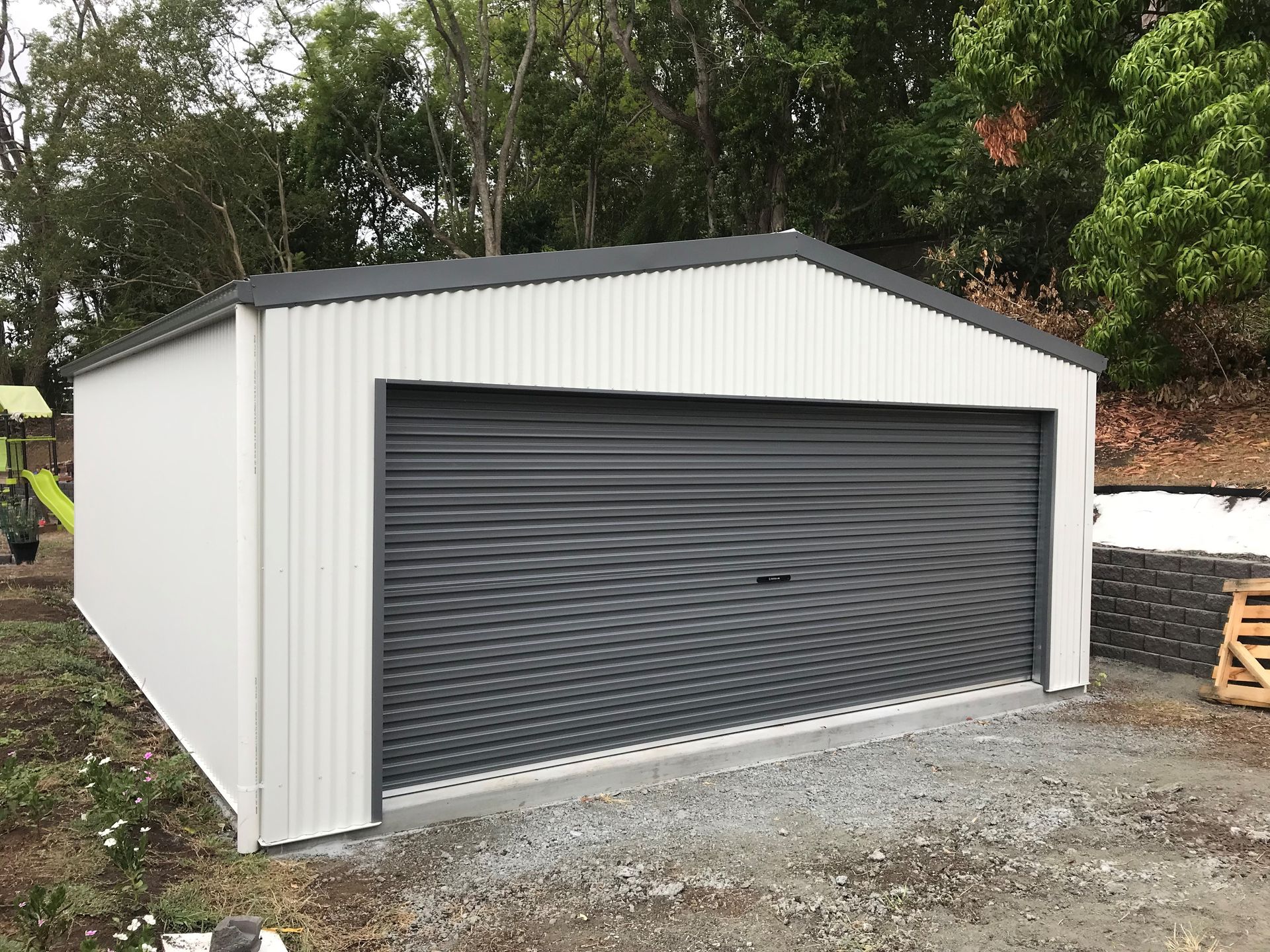 White and grey metal garage with a roller door, on a gravel base, set against a backdrop of trees - Northern Rivers Sheds in South Lismore, NSW