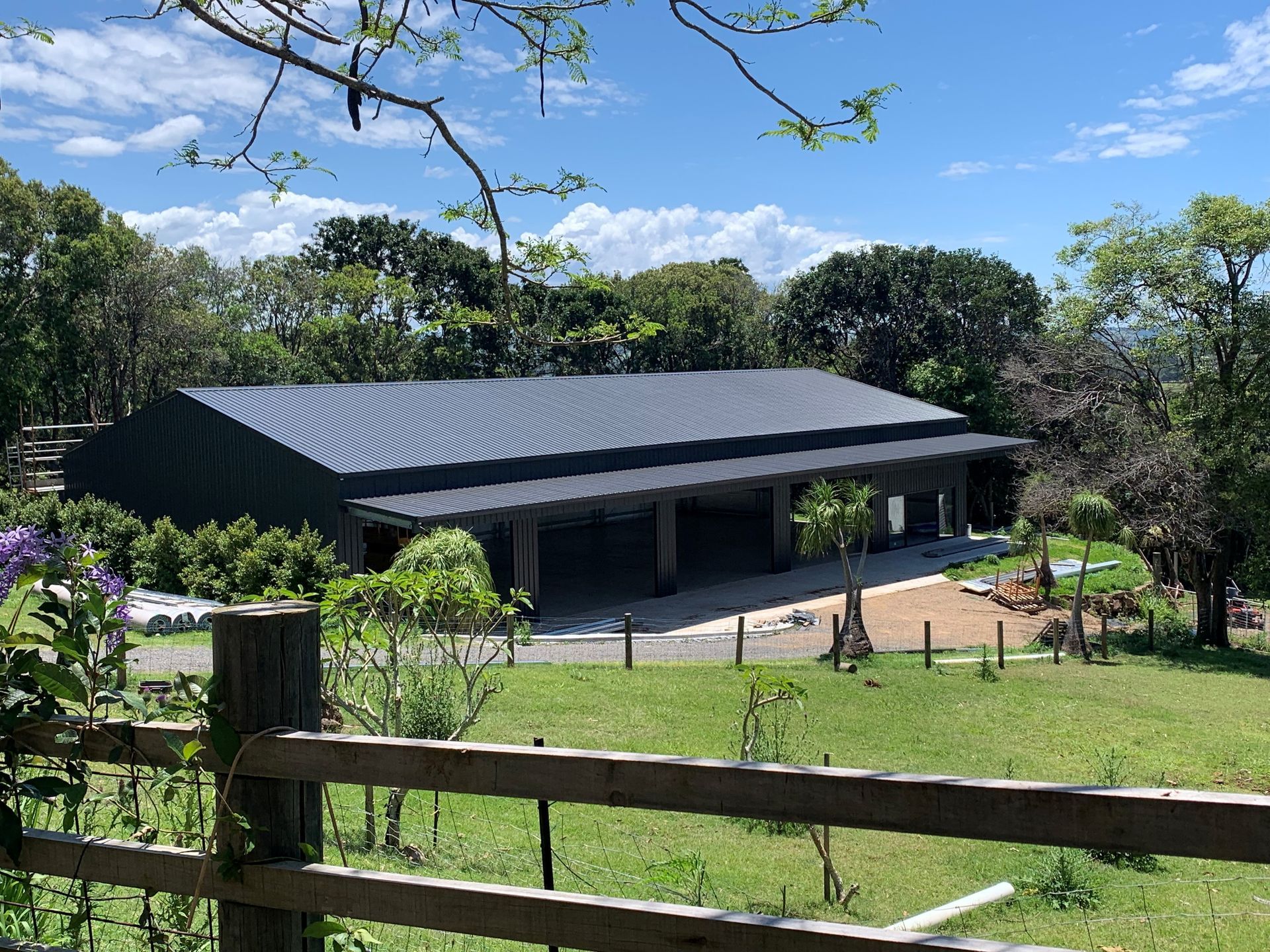 Building with dark roof and open-air structure, set in a grassy landscape with trees and a blue sky— Northern Rivers Sheds in Byron Bay, NSW
