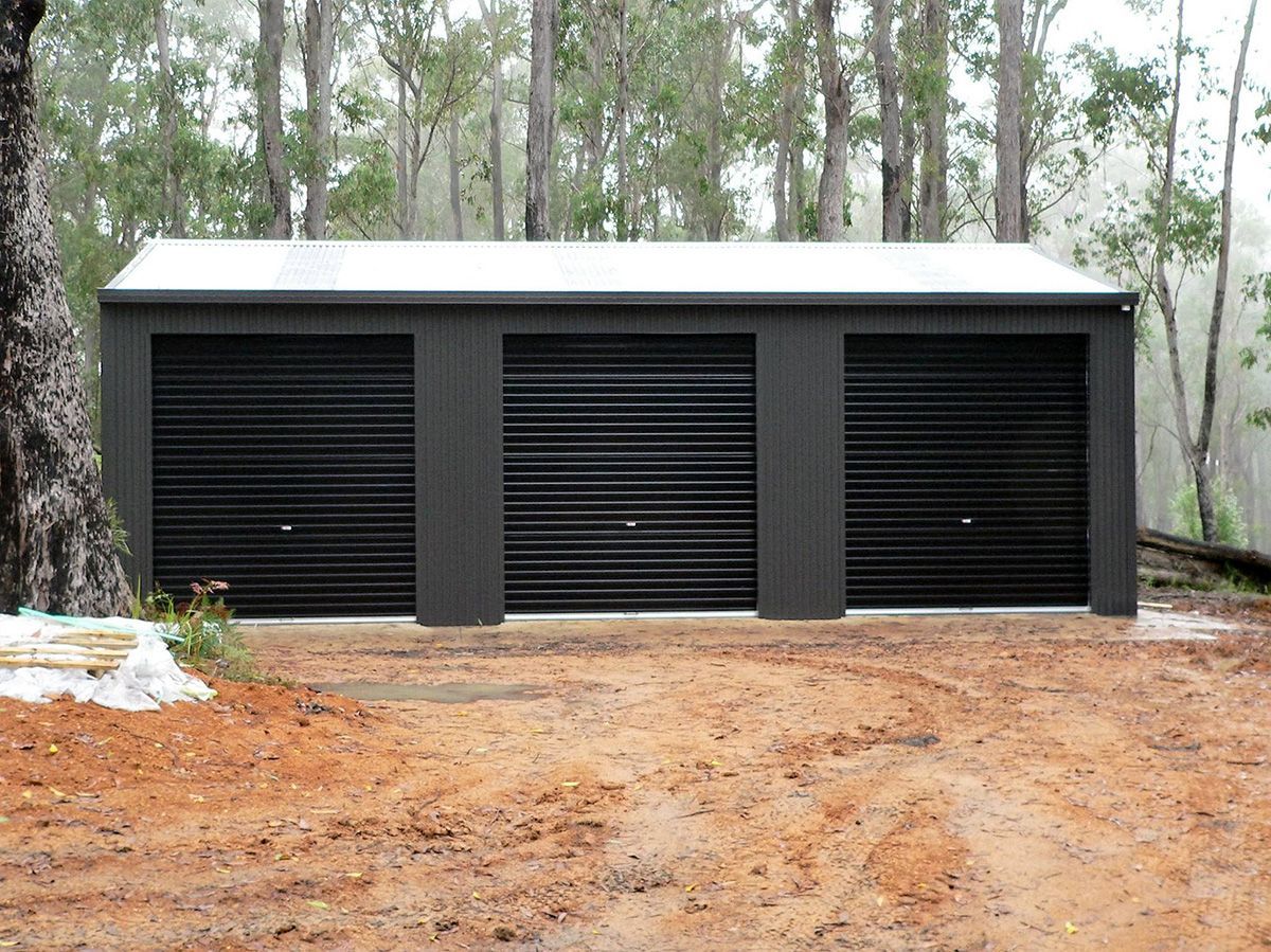 Dark gray three-car garage with black roll-up doors, set in a forest clearing.