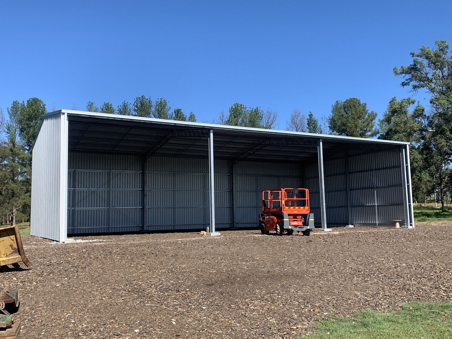 Open-sided metal shed on gravel ground, orange lift inside, trees in background, sunny day - Northern Rivers Sheds in South Lismore, NSW