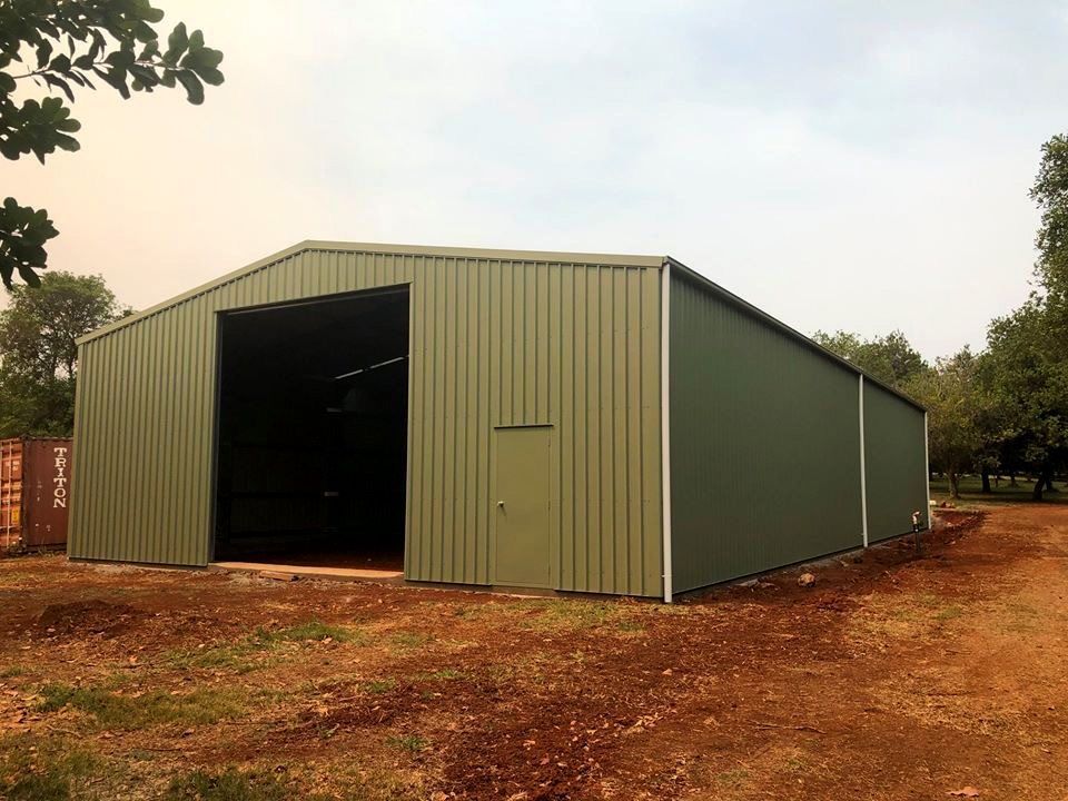 Green metal shed with large opening, small door, set on a dirt field with trees in the background.