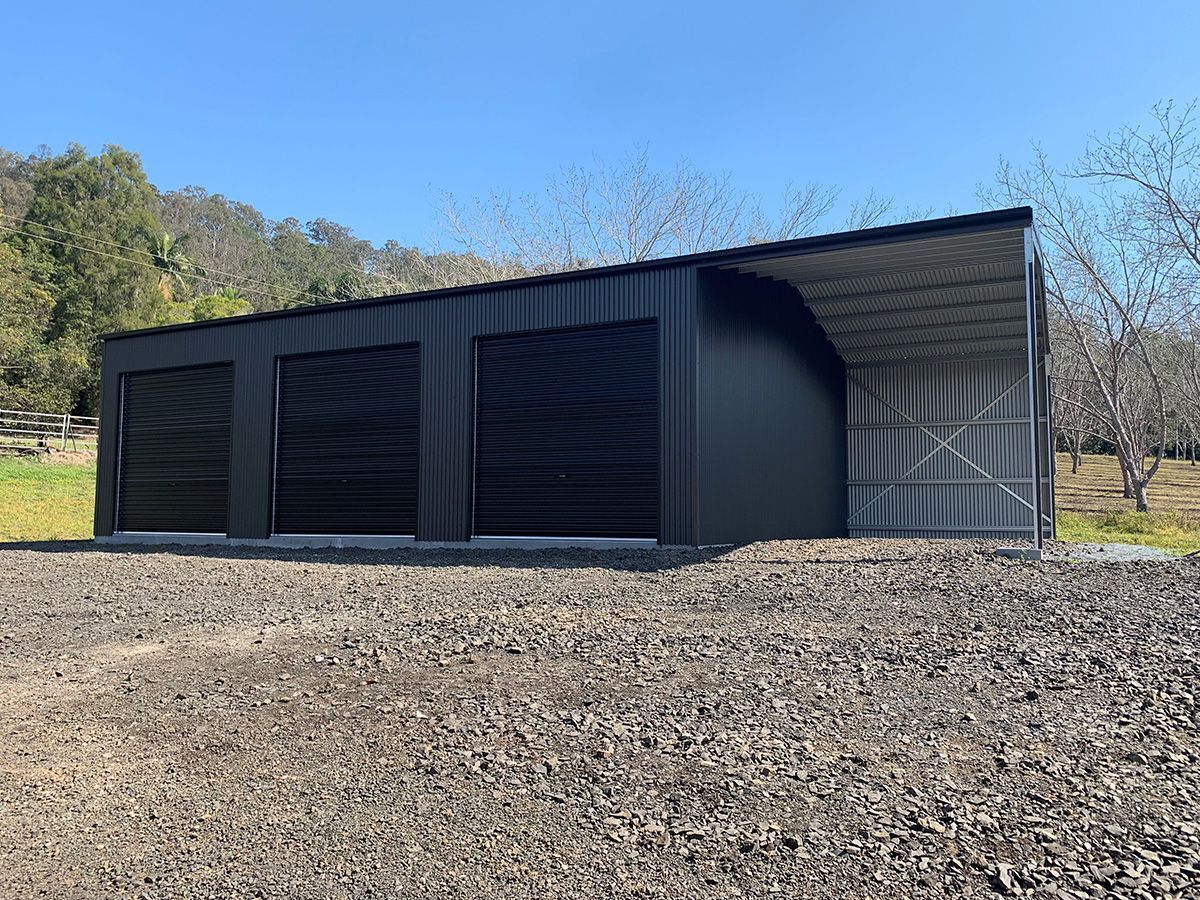 Black metal storage building with three garage doors and a mesh awning, set on gravel.