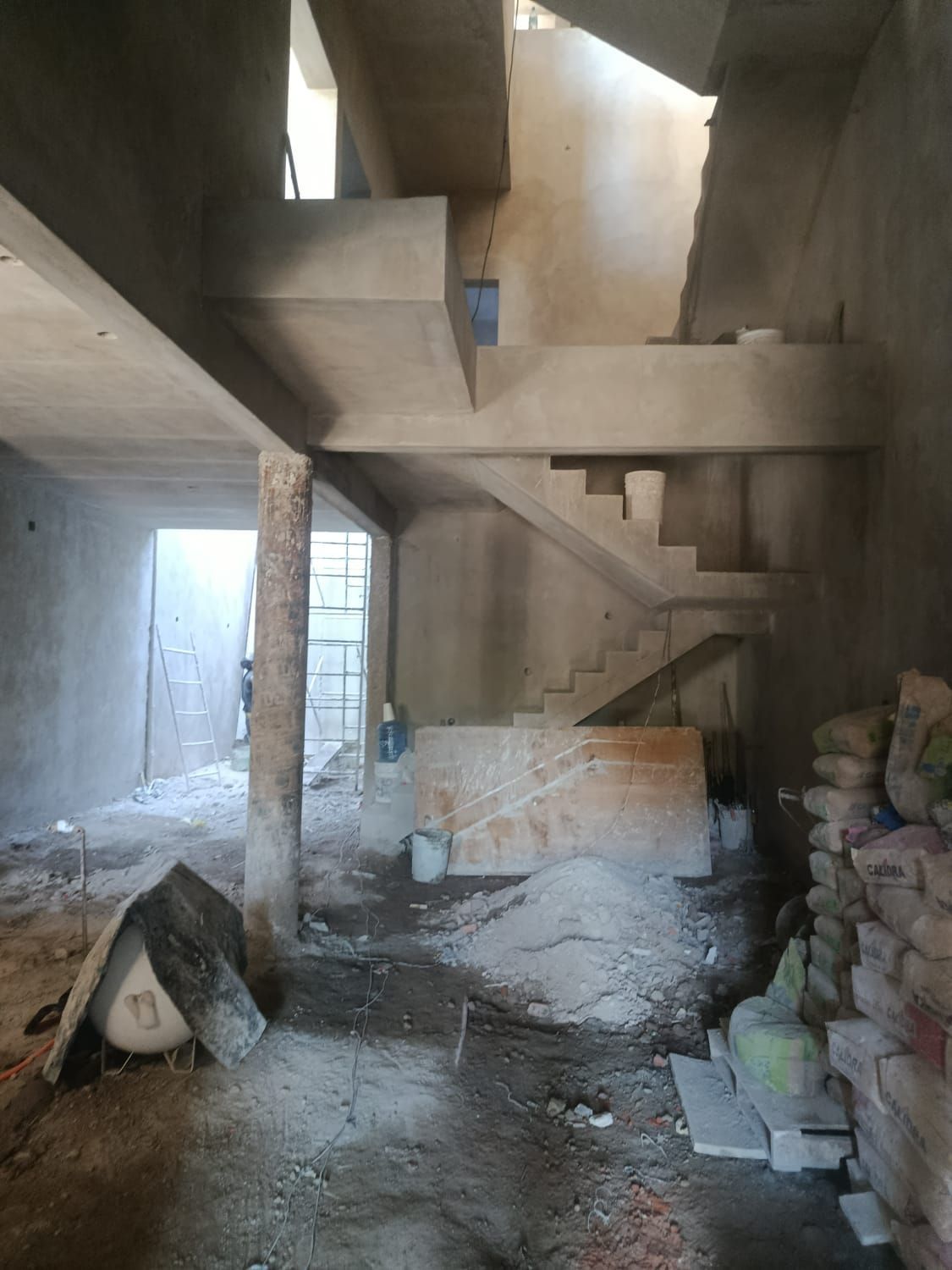 Interior de una habitación en construcción con escaleras de hormigón, un pilar de soporte y materiales de construcción dispersos.