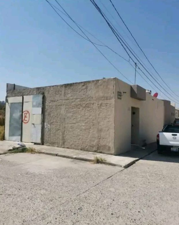 Un edificio de color beige, con techo plano, puertas metálicas y un cartel visible de prohibido aparcar, en un día soleado, junto a un camión blanco estacionado.