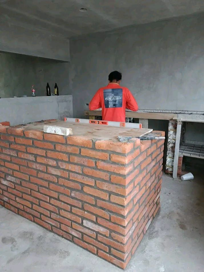 Un obrero de la construcción con una camisa naranja está construyendo una isla de cocina de ladrillo en una habitación sin terminar con paredes de hormigón.