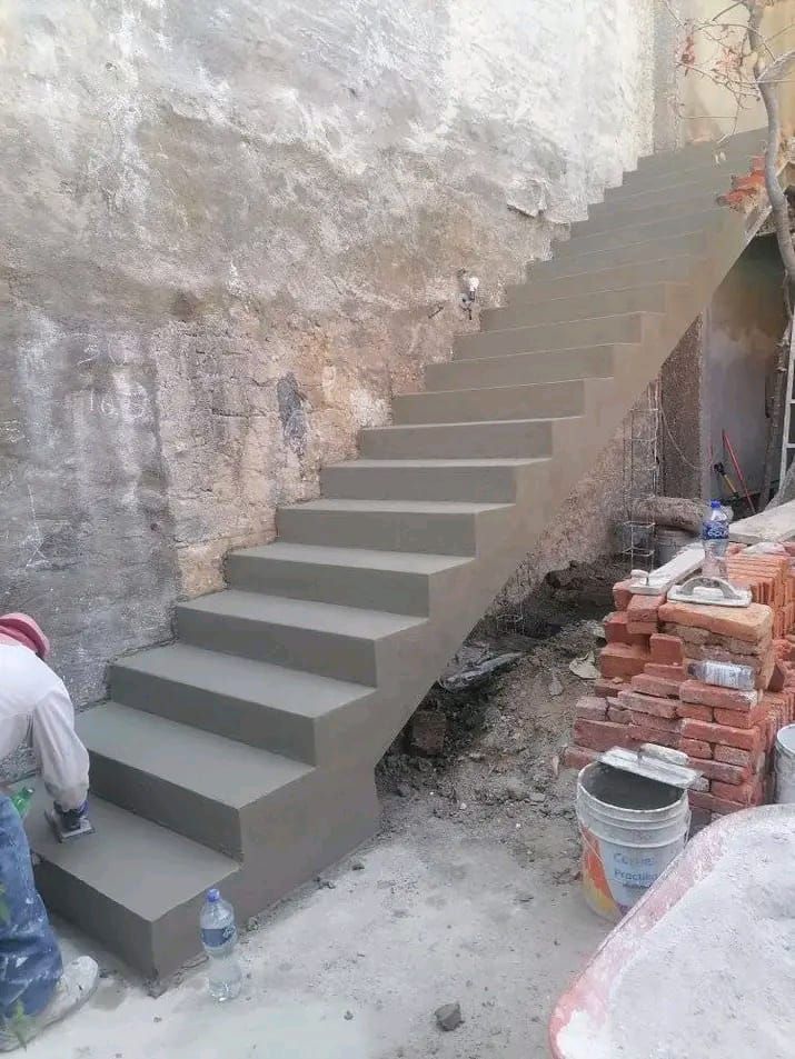 Un obrero de la construcción termina de construir una escalera de hormigón recién vertido junto a un muro de piedra irregular.