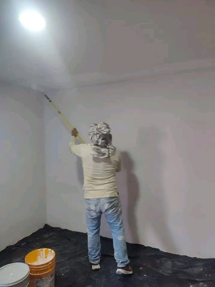 Una persona que lleva la cabeza cubierta y ropa informal utiliza un palo extensible para pintar el techo blanco de una habitación.