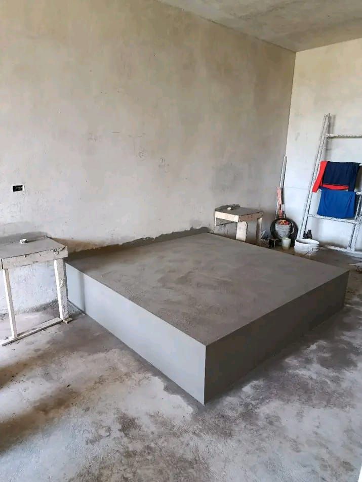 Una estructura de plataforma de hormigón situada en el centro de una habitación con paredes sin terminar, cerca de un estante metálico que contiene mantas dobladas.