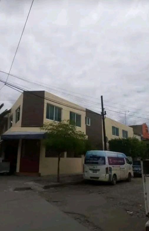 Un edificio de dos plantas de color beige con detalles marrones se alza en una calle con una furgoneta aparcada delante, bajo un cielo nublado.