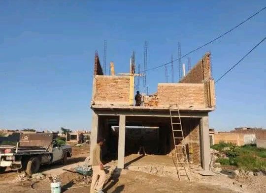 Los trabajadores construyen una estructura de ladrillo de dos pisos bajo un cielo azul despejado, con un camión estacionado cerca en un terreno baldío.
