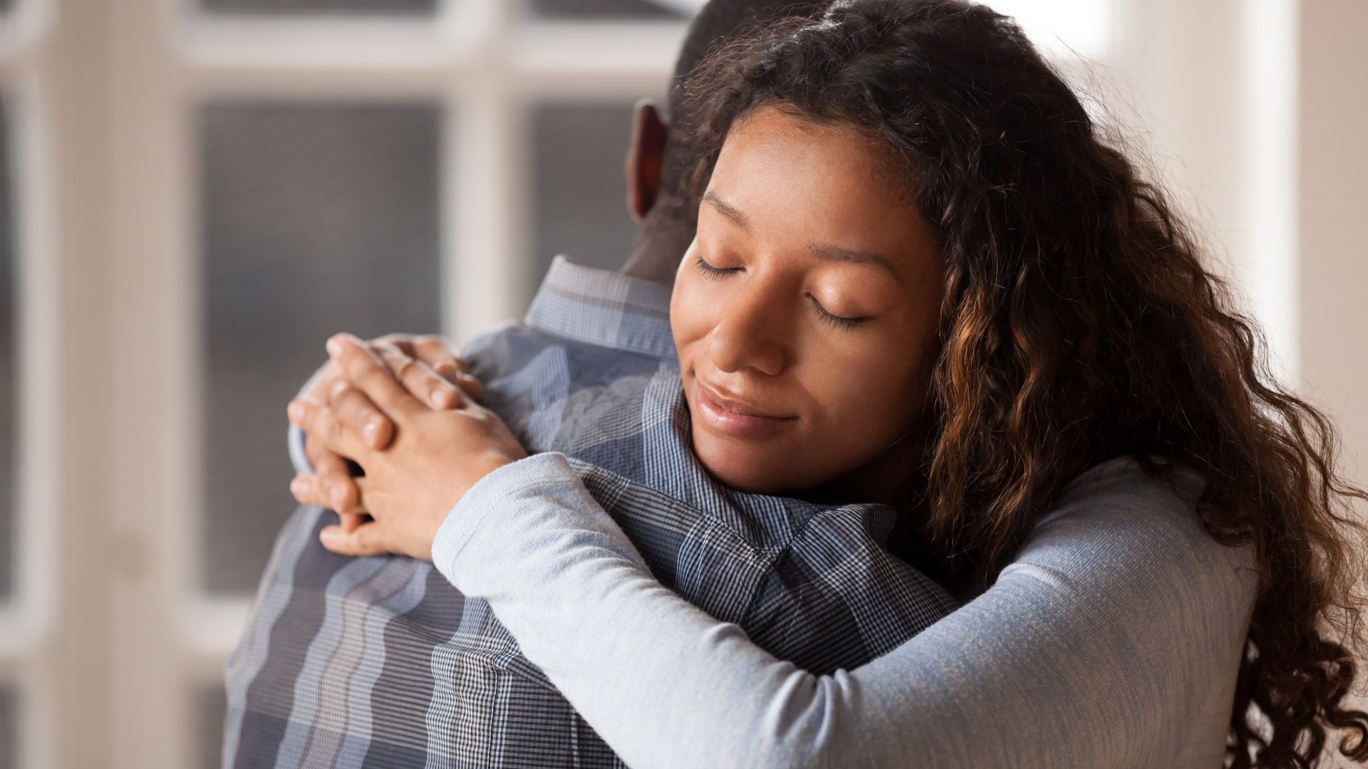 Two people hugging tightly indoors, one with eyes closed and a calm expression