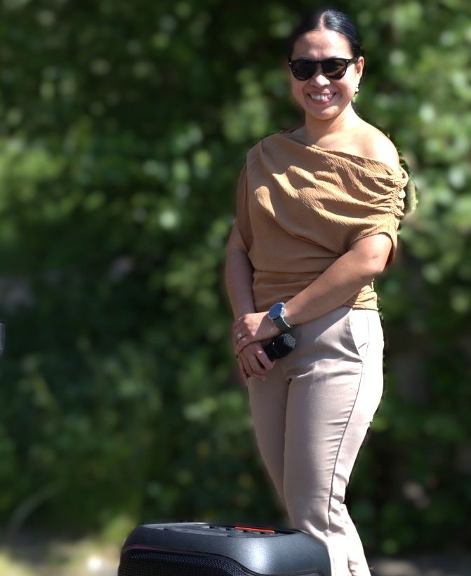 Woman in tan top and pants smiles, stands by a black object against a blurred green backdrop.