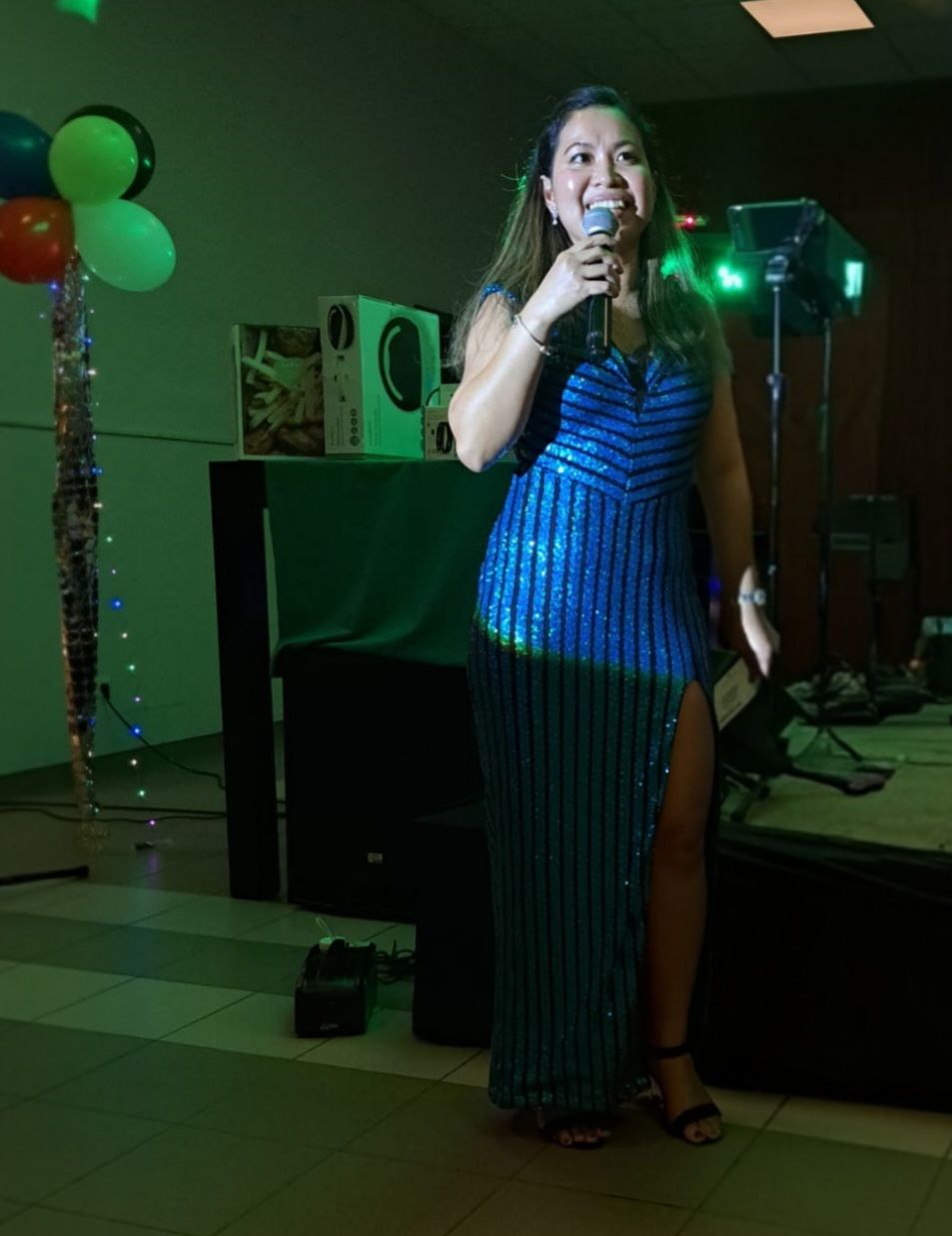 Woman in a blue sequined gown singing into a microphone at an event, with balloons and speakers in the background.