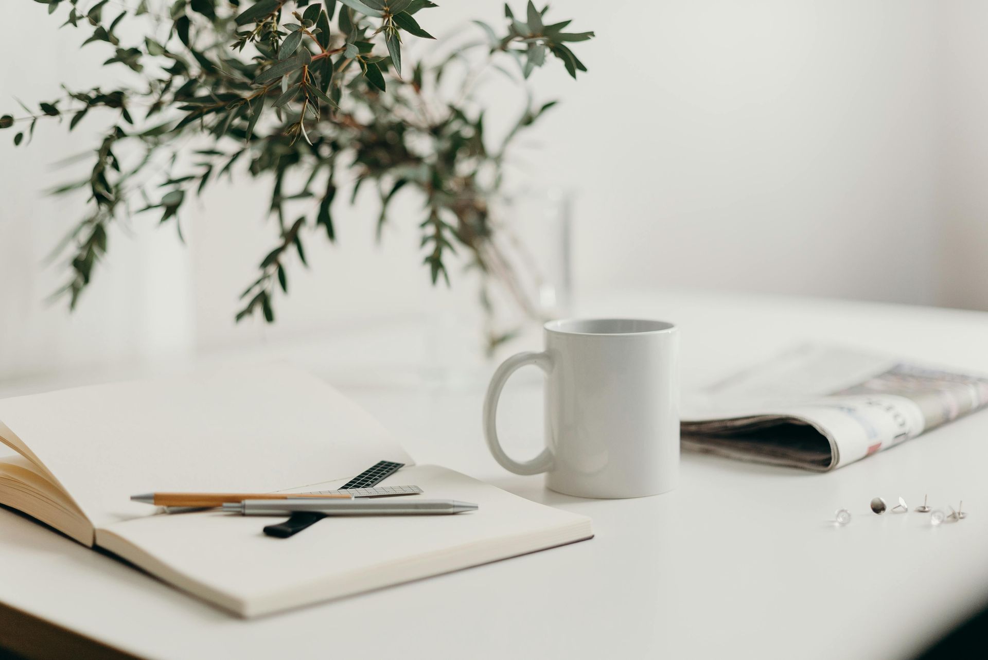 White table with a mug, open notebook, pens, newspaper, and vase of greenery.