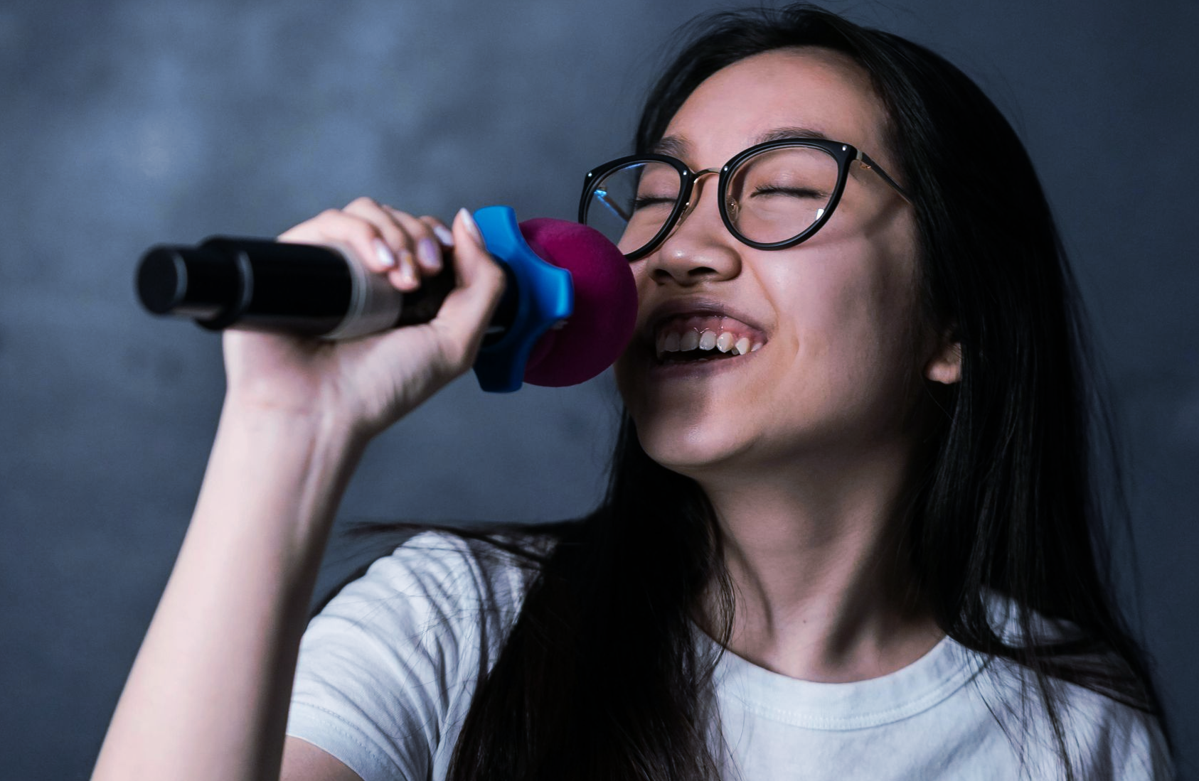Woman with glasses singing into a microphone, eyes closed, smiling, against a gray background.
