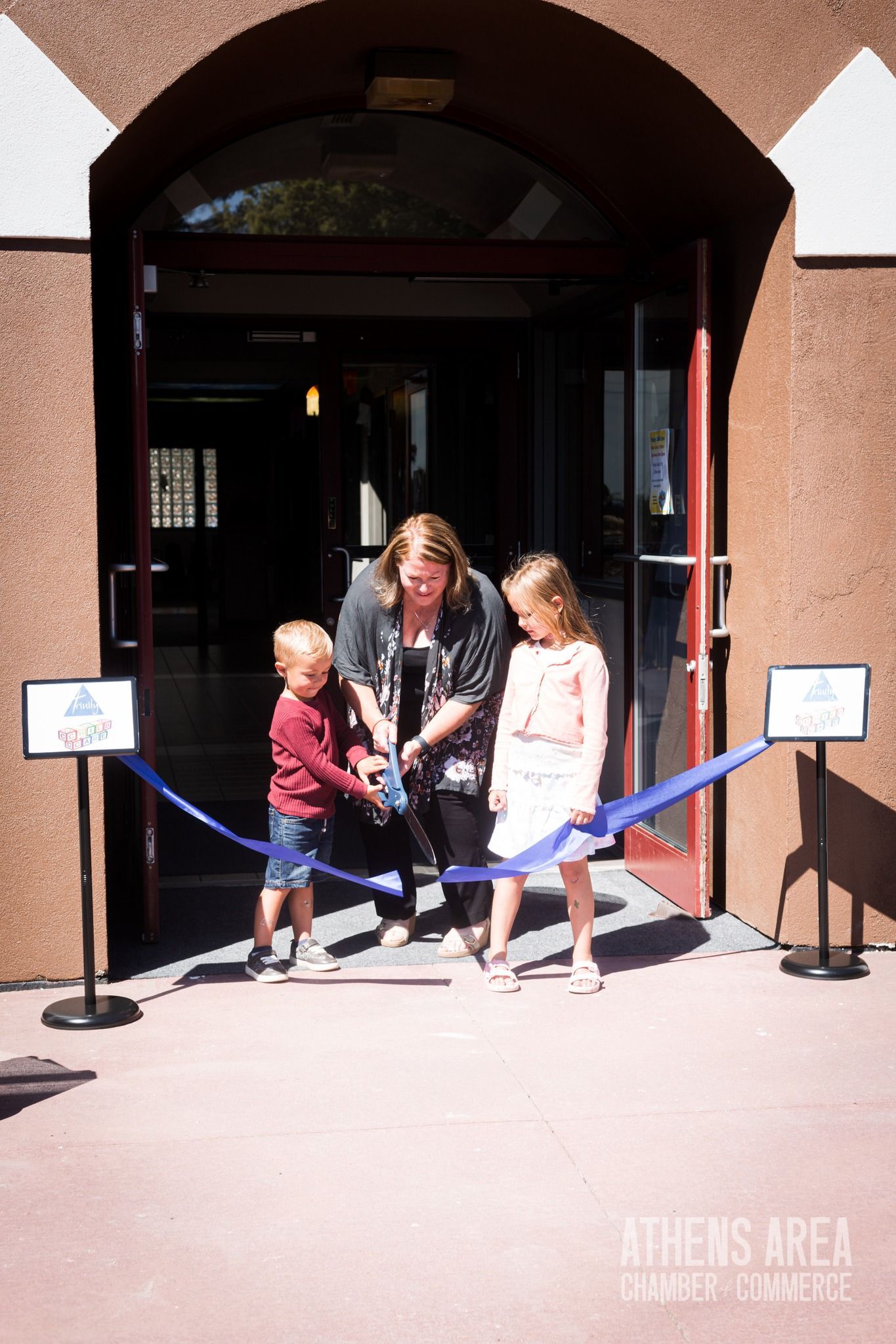 A woman and two children are cutting a blue ribbon in front of a building.