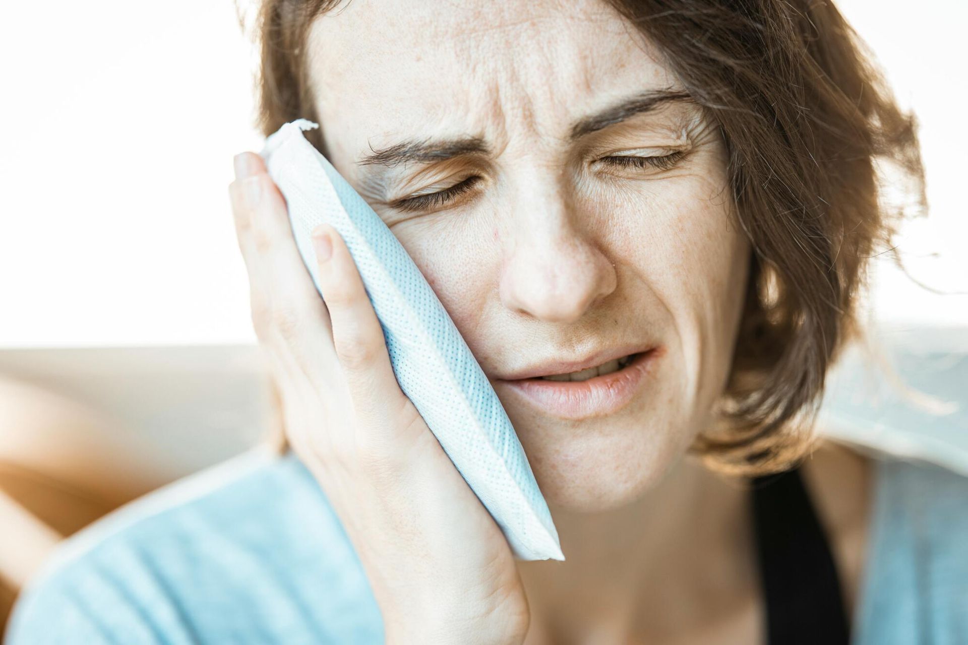 Woman holding a cold compress to her cheek, grimacing, suggesting tooth pain.