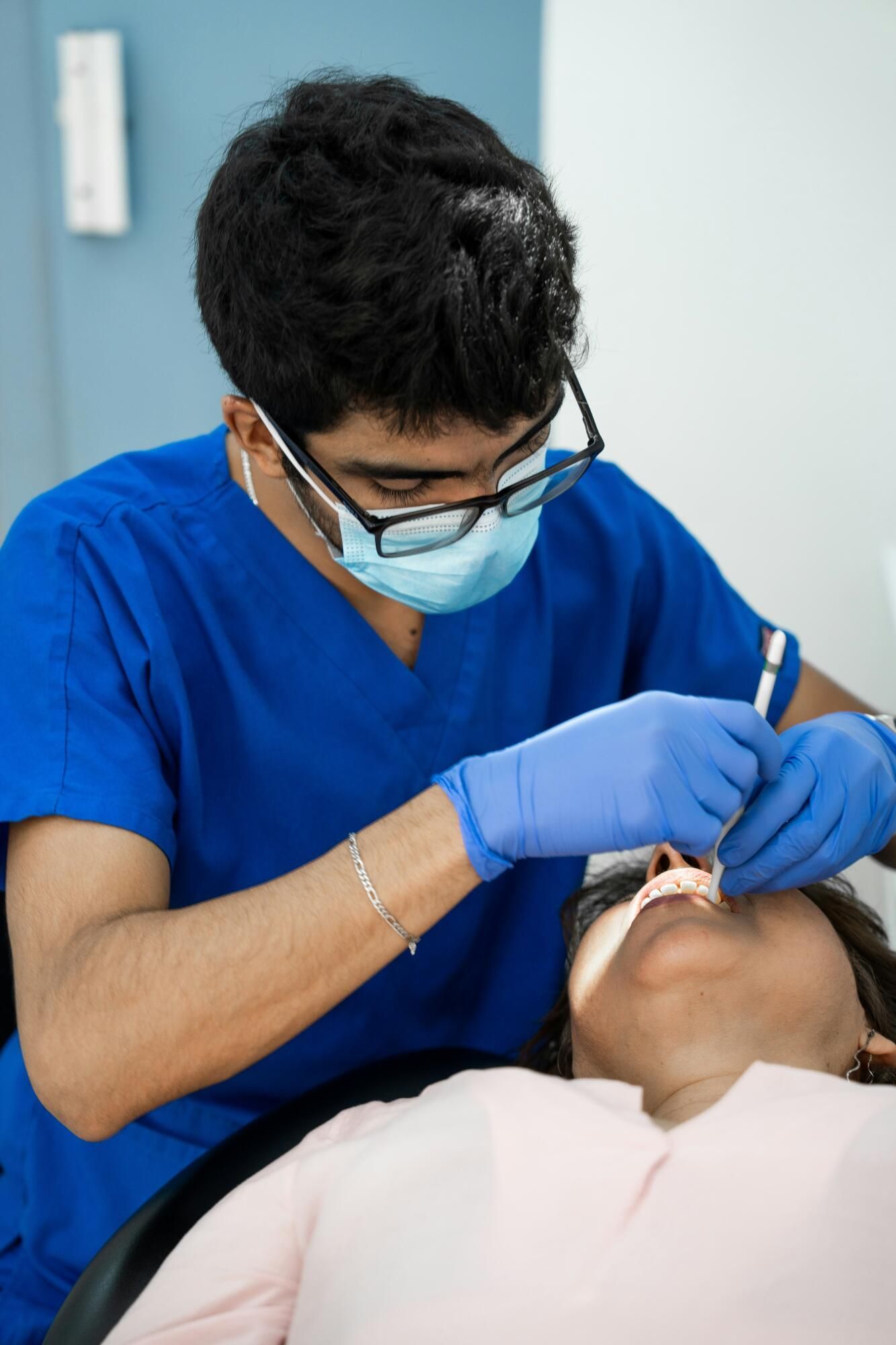 A dental professional in blue scrubs, mask, and gloves performs a routine examination on a patient in a dental chair.